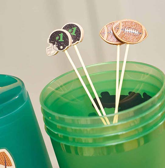 The Superbowl Set by Sophistiplate features four wooden drink stirrers with football and helmet designs in a green plastic cup, perfect for pairing with party-in-a-box sets or designer disposable plates. A second cup appears partially on the left.