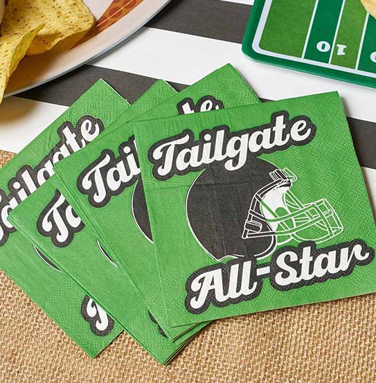 The Superbowl Set by Sophistiplate features green napkins with "Tailgate All-Star" and a football helmet, displayed on a striped tablecloth near disposable plates of chips and a football field-themed tray.
