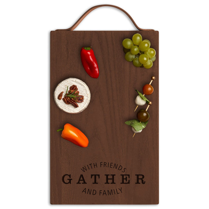 The Maple Thermal Ash Plank 16x10" Cheese & Charcuterie Board features “Gather with Friends & Family” engraving, a leather handle, and is shown with cheese, pecans, peppers, green grapes, and skewers. Made in the USA.