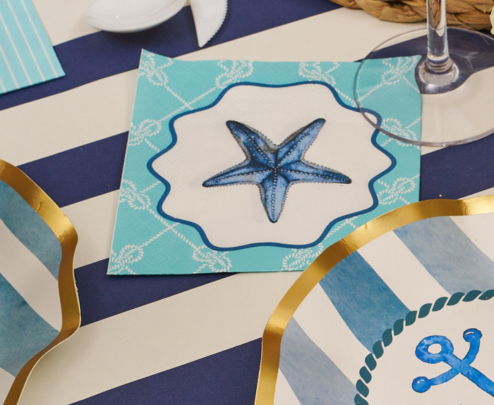 The Tidal Wave Table Setting by Sophistiplate features a blue and white starfish napkin, a navy striped tablecloth, and disposable plates with gold edges and anchor motifs—ideal for easy coastal party decor.