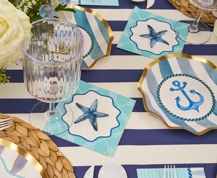 Sophistiplate’s Tidal Wave Table Setting includes blue and white striped plates, starfish and anchor motifs, gold accents, a clear goblet, and woven placemats—making coastal party decor easy and stylish.