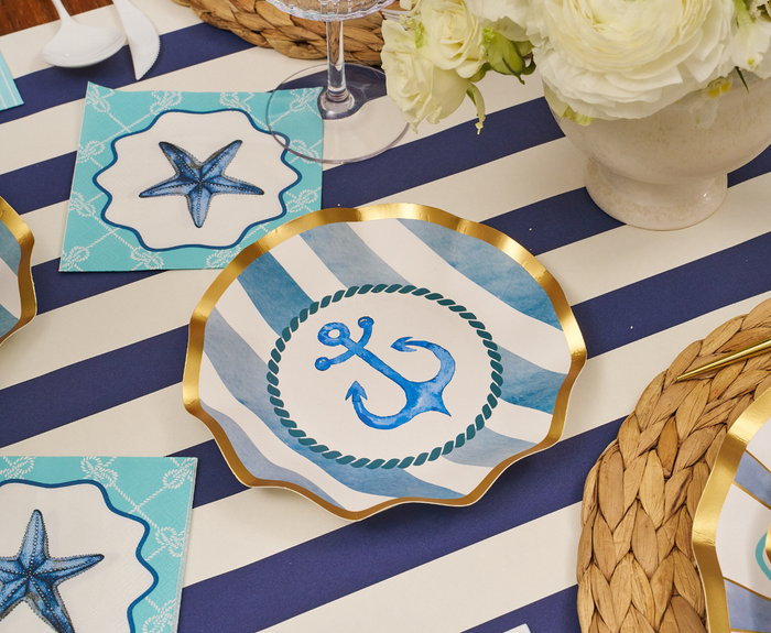 The Tidal Wave Table Setting by Sophistiplate features a scallop-shaped plate with a blue anchor and gold trim on a navy and white striped tablecloth, accented by starfish napkins, a woven placemat, and white flowers for coastal flair.
