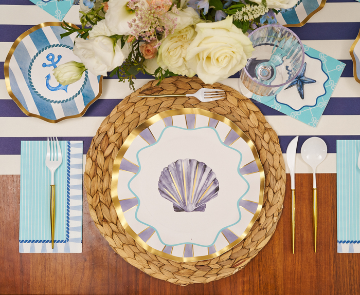 The Tidal Wave Table Setting by Sophistiplate includes a shell plate, woven placemat, blue-striped tablecloth, anchor plate, floral centerpiece, gold utensils, blue marine-themed napkin, and clear cup for a nautical tablescape.