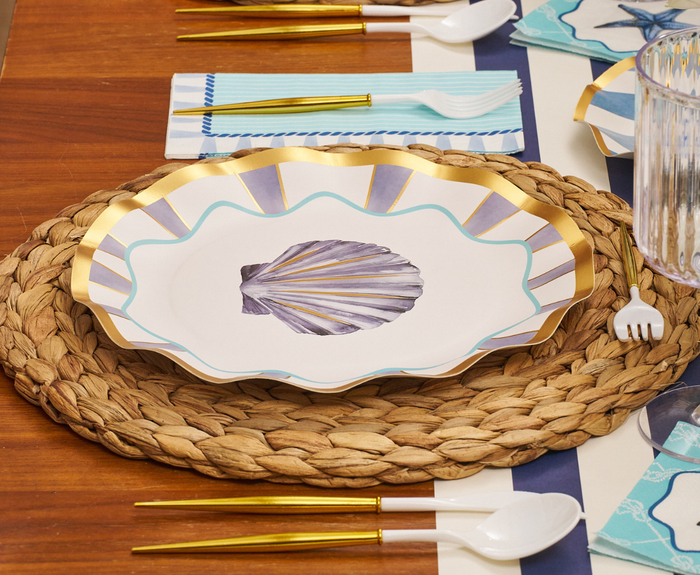 The Tidal Wave Table Setting by Sophistiplate features a scallop shell plate with gold and blue accents, gold and white cutlery, striped napkins, a clear ribbed glass, and a woven placemat for coastal-inspired decor.