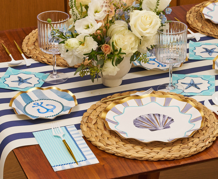 The Tidal Wave Table Setting by Sophistiplate features shell-and-anchor disposable plates, woven placemats, a striped runner, blue-striped napkins, gold cutlery, glassware, and a white floral centerpiece for chic coastal style.