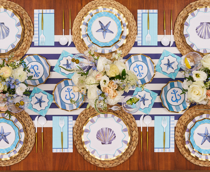 The Tidal Wave Table Setting by Sophistiplate features coastal party decor for six, including woven placemats, nautical disposable plates, blue-striped napkins, gold cutlery, and floral centerpieces with white and blush flowers.