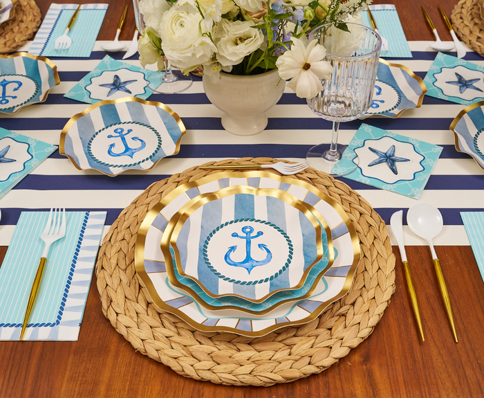 The Tidal Wave Table Setting by Sophistiplate features anchor plates, blue-striped placemats, gold utensils, starfish napkins, and a white floral centerpiece—ideal coastal party decor for any wooden table.