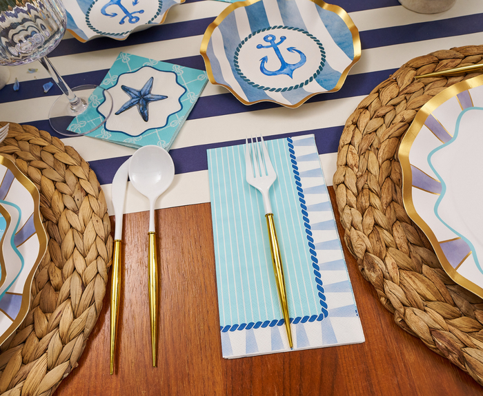 The Tidal Wave Table Setting by Sophistiplate includes anchor and starfish plates and napkins, a blue-and-white striped runner, woven placemats, and gold-handled cutlery—everything for the perfect coastal tablescape.