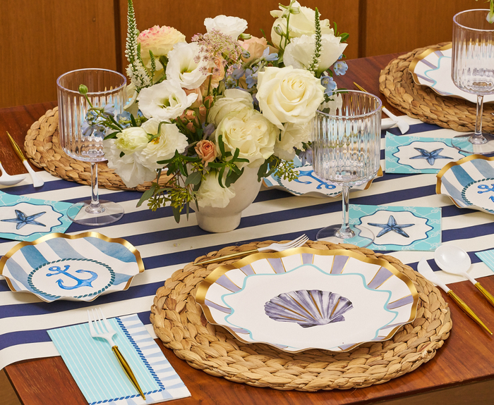 The Tidal Wave Table Setting by Sophistiplate features shell and anchor disposable plates, woven placemats, striped napkins, gold flatware, clear glasses, and a centerpiece of white and pastel flowers for coastal party decor.