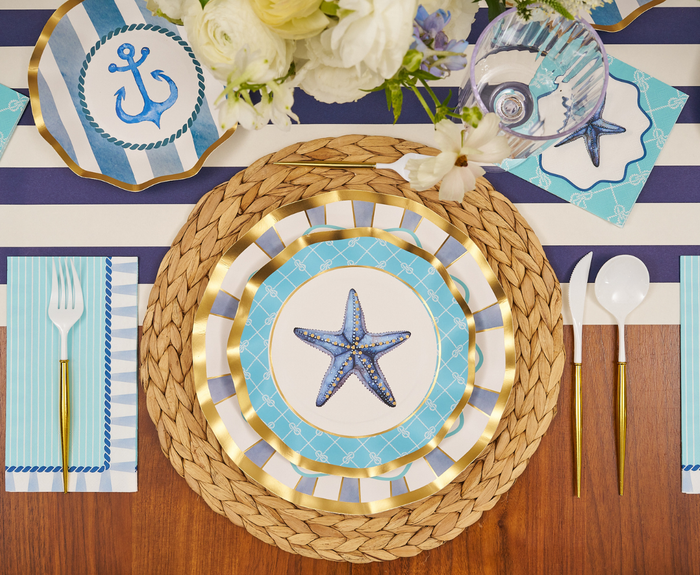 The Tidal Wave Table Setting by Sophistiplate includes disposable starfish plates, striped napkins, gold utensils, a woven placemat, and a white floral centerpiece—an easy nautical-themed tablescape with blue, white, and gold accents for your coastal party.