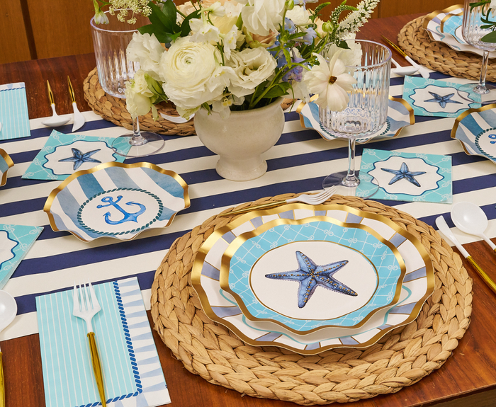 The Tidal Wave Table Setting by Sophistiplate is a nautical-themed party decor set with woven placemats, a blue and white striped runner, starfish and anchor disposable plates, gold cutlery, glassware, vase with white flowers, and blue-striped napkins.
