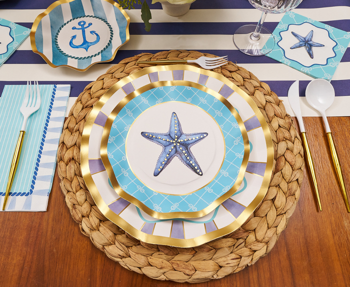 Create a stunning nautical vibe with the Tidal Wave Table Setting by Sophistiplate—includes a woven placemat, blue and white starfish plates, gold utensils, and anchor-striped napkins for stylish entertaining.