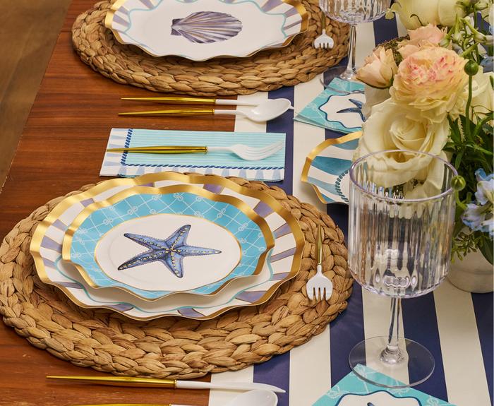 The Tidal Wave Table Setting from Sophistiplate features woven placemats, striped starfish-and-seashell plates, gold and white cutlery, striped napkins, clear glasses, and a white-and-yellow floral centerpiece for perfect coastal party decor.