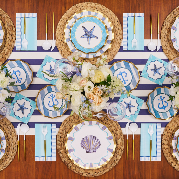 Create a nautical-themed tablescape with the Sophistiplate Tidal Wave Table Setting, featuring seashell, starfish, and anchor motifs, coordinating blue-and-white accents, gold utensils, and woven placemats.