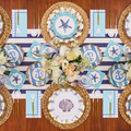 Create a nautical-themed tablescape with the Sophistiplate Tidal Wave Table Setting, featuring seashell, starfish, and anchor motifs, coordinating blue-and-white accents, gold utensils, and woven placemats.
