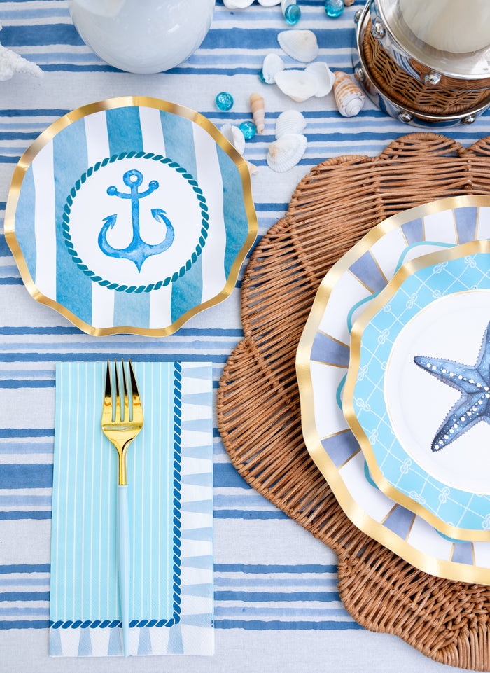 The Sophistiplate Tidal Wave Table Setting features blue and white striped plates with anchor and starfish designs, a gold fork on a striped blue napkin, wicker placemats, and seashell accents for a nautical-inspired party look.