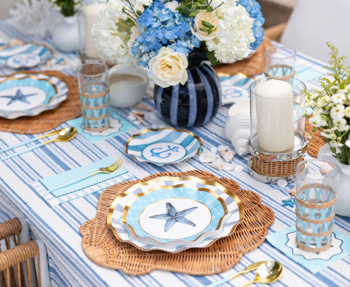 Create a coastal-themed party table with Sophistiplate's Tidal Wave Table Setting, featuring blue and white striped linens, starfish paper plates, wicker placemats, gold utensils, floral centerpieces, candles, and seashells.