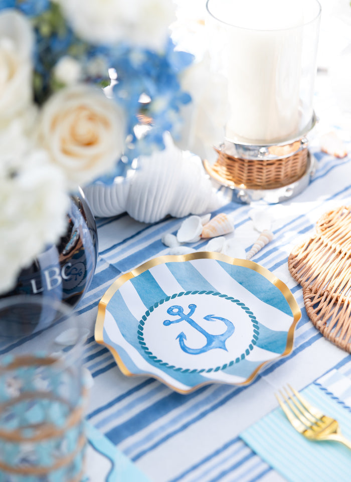 Create a nautical-themed party with the Sophistiplate Tidal Wave Table Setting, featuring coastal disposable plates, blue and white linens, wicker accents, seashells, fresh flowers, and a glass candle holder.