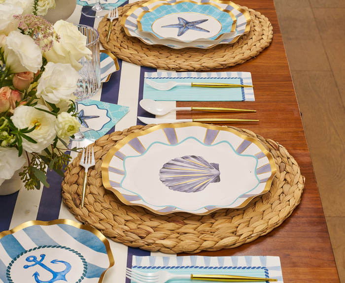 The Tidal Wave Table Setting by Sophistiplate features woven placemats, seashell and starfish plates, gold cutlery, striped napkins, and white flowers for a stylish coastal party tablescape.