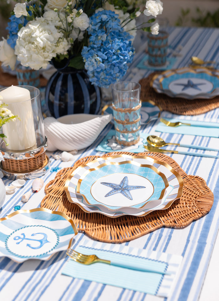 Create a coastal-themed tablescape with the Tidal Wave Table Setting by Sophistiplate, featuring designer starfish and anchor plates, blue-and-white accents, gold cutlery, wicker placemats, seashells, and a blue-and-white floral centerpiece.