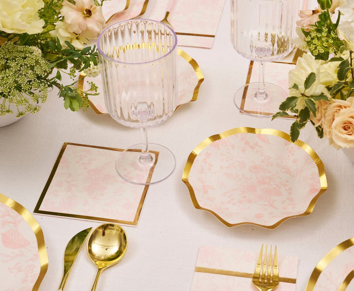 Create a stunning tablescape with the Sophistiplate Timeless Table Setting: Blush—featuring blush and gold plates, gold utensils, ribbed glassware, floral napkins, and flower accents on a white cloth—perfect for any celebration.