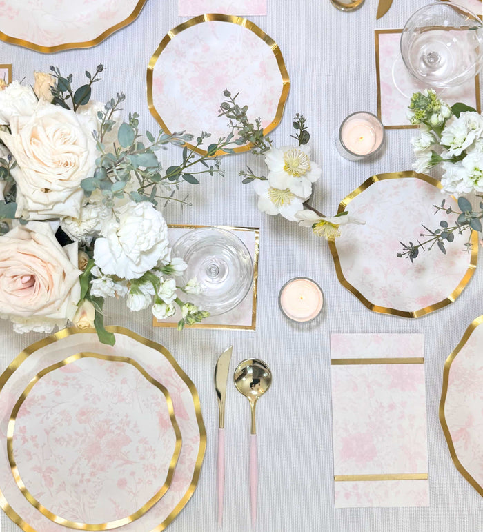 Elegant table setting with Sophistiplate Timeless Blush Salad Plates, matching napkins, gold flatware, white and blush flowers, candles, and wine glasses on a textured white tablecloth.