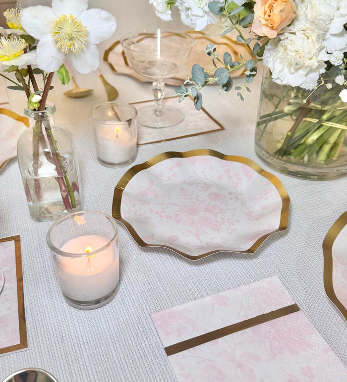 The Timeless Table Setting: Blush by Sophistiplate features elegant paper plates with gold edges, floral place cards, glass candle holders, and peach-and-white florals on a textured cloth for a refined party-in-a-box look.