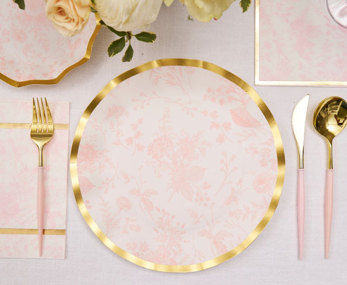Sophistiplate Timeless Blush Dinner Plates (8pk) in pastel pink with a gold foil edge are arranged on a white tablecloth, paired with gold and pink cutlery, a matching napkin, and a floral centerpiece.