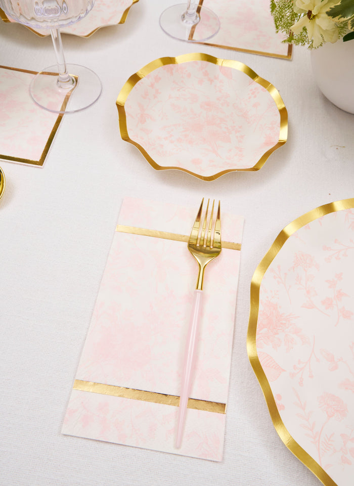 The Timeless Table Setting: Blush by Sophistiplate features scalloped, floral-patterned pink and gold heavy-duty paper plates, gold-edged napkins, pink-handled gold forks, and matching glassware on a white tablecloth.