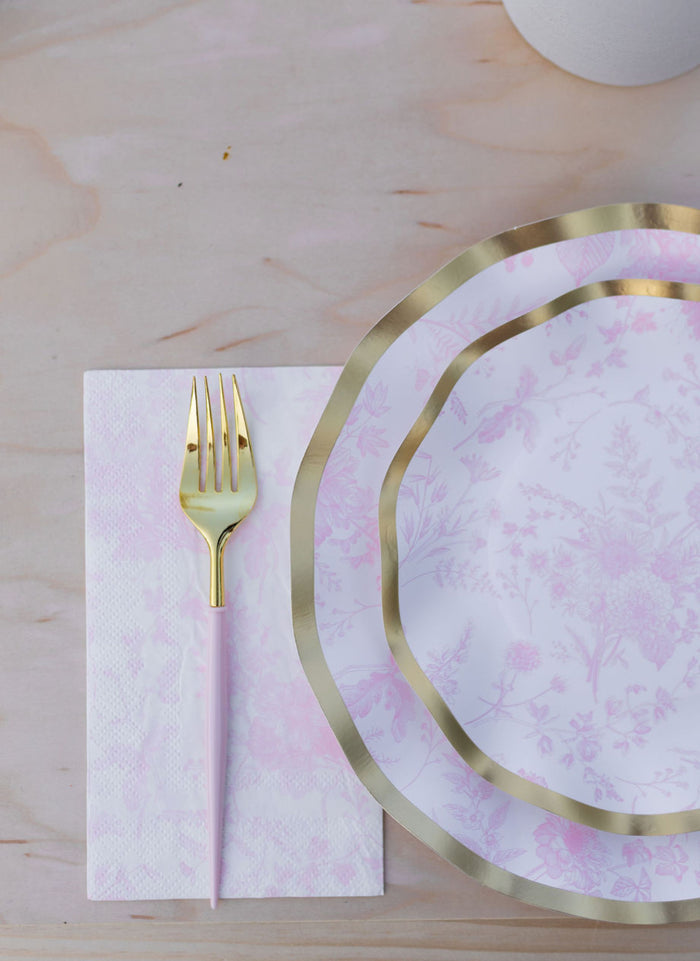 The Sophistiplate Timeless Table Setting: Blush features a gold and white fork resting on a floral napkin beside two pink and white heavy duty paper plates with gold rims, all arranged on a light wooden table.