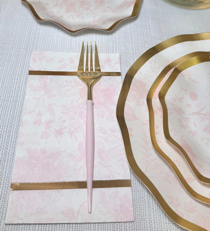 A Sophistiplate Timeless Blush Guest Towel rests on a textured white tablecloth, beside matching pink and gold-rimmed plates and elegant gold-accented flatware. Pack includes 20 guest towels.