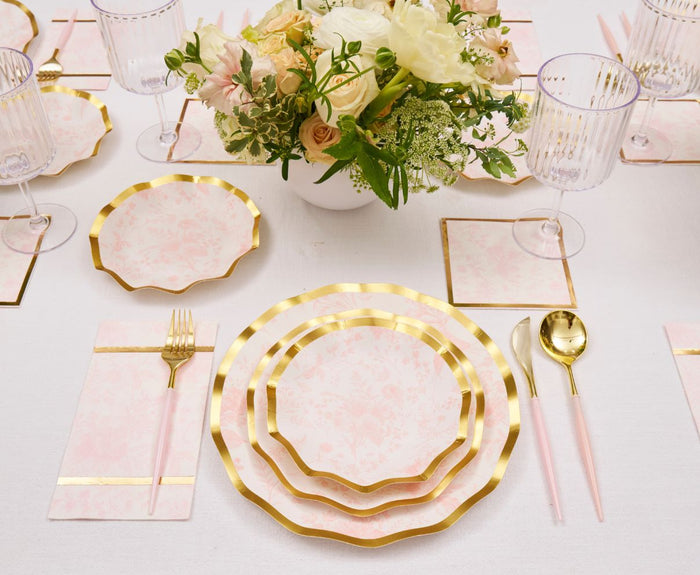 An elegant table is set with gold-rimmed plates, utensils, striped glasses, a cream and blush floral centerpiece, and Timeless Blush Cocktail Napkins by Sophistiplate adding a charming touch to the white tablecloth.