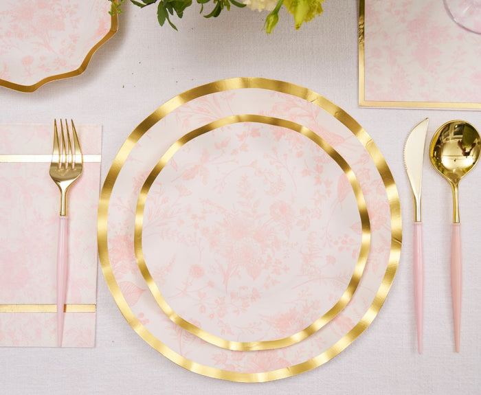 A table is set with Sophistiplate's Timeless Blush Salad Plates, featuring pink florals and gold edges, complemented by pink-handled gold cutlery and a matching napkin on a white tablecloth. A floral arrangement is partly visible above.