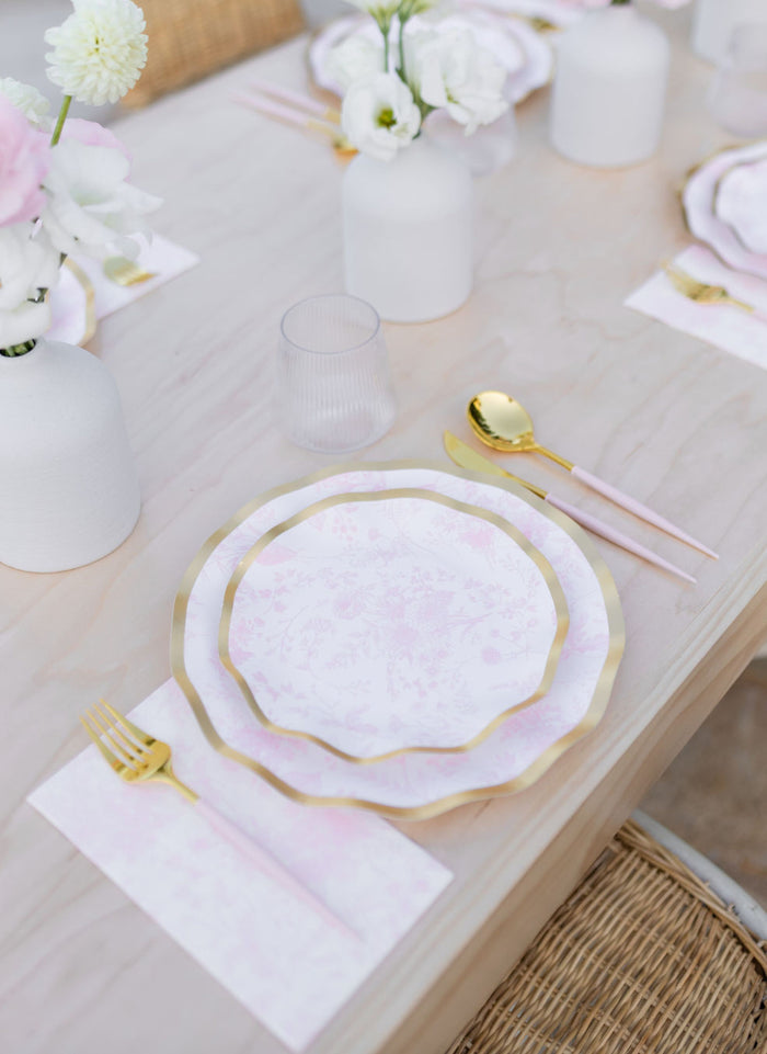 Elegant table setting with Sophistiplate Timeless Blush Salad Plates (8pk), pink-handled gold cutlery, a ribbed glass, and white floral arrangements in matte vases on a light wood table with woven chairs.