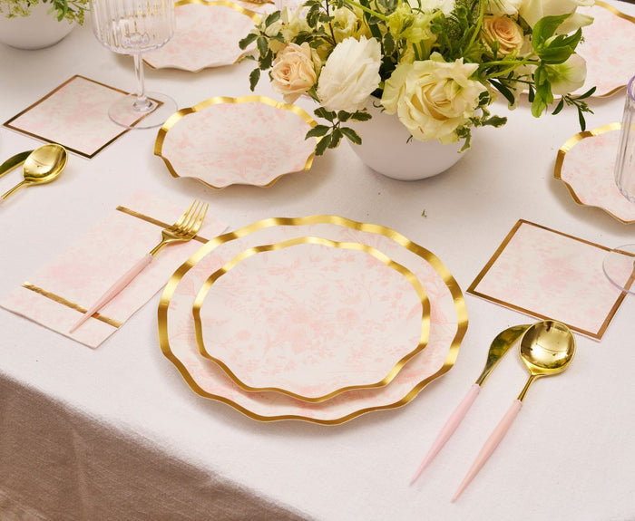 A table is set with Sophistiplate Timeless Blush Salad Plates (8pk), matching cutlery, white floral centerpieces, glassware, napkins, and gold-edged place cards—all on a beige tablecloth for an elegant touch.