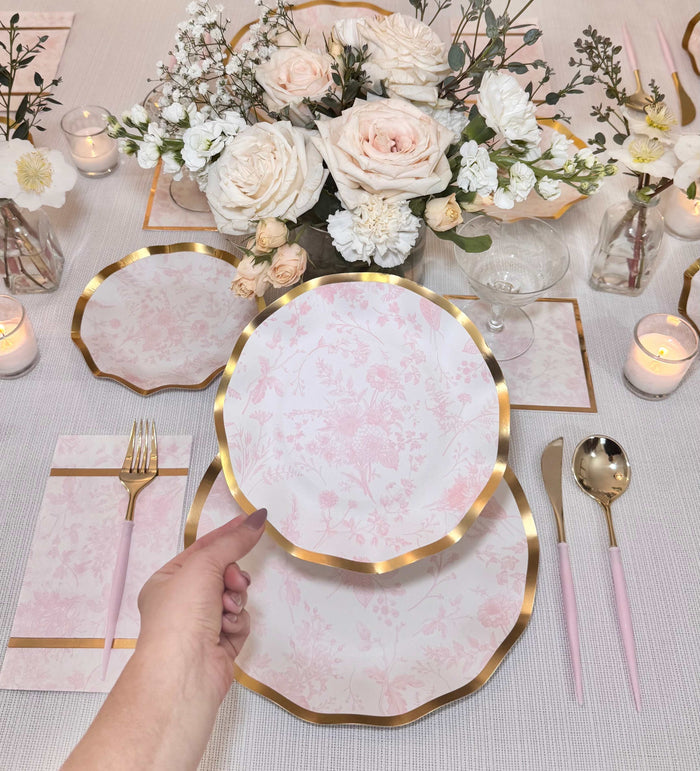 A hand holds a Sophistiplate Timeless Blush Salad Plate from an 8-pack above a matching table setting with pink and gold dishes, cutlery, napkins, blush and white florals, and candles for an elegant look.