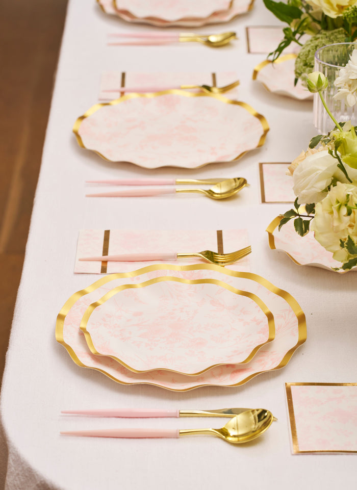 Create a chic tablescape with the Timeless Table Setting: Blush by Sophistiplate, featuring scalloped pink and gold disposable plates, pink-handled cutlery, white napkins with gold accents, and cream floral centerpieces on a white tablecloth.