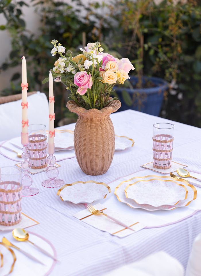 A white outdoor table features Sophistiplate Timeless Blush Salad Plates, gold flatware, pink glasses, and two tall white candles with pink bows. Pastel blooms in a woven vase create a centerpiece, surrounded by lush green plants.