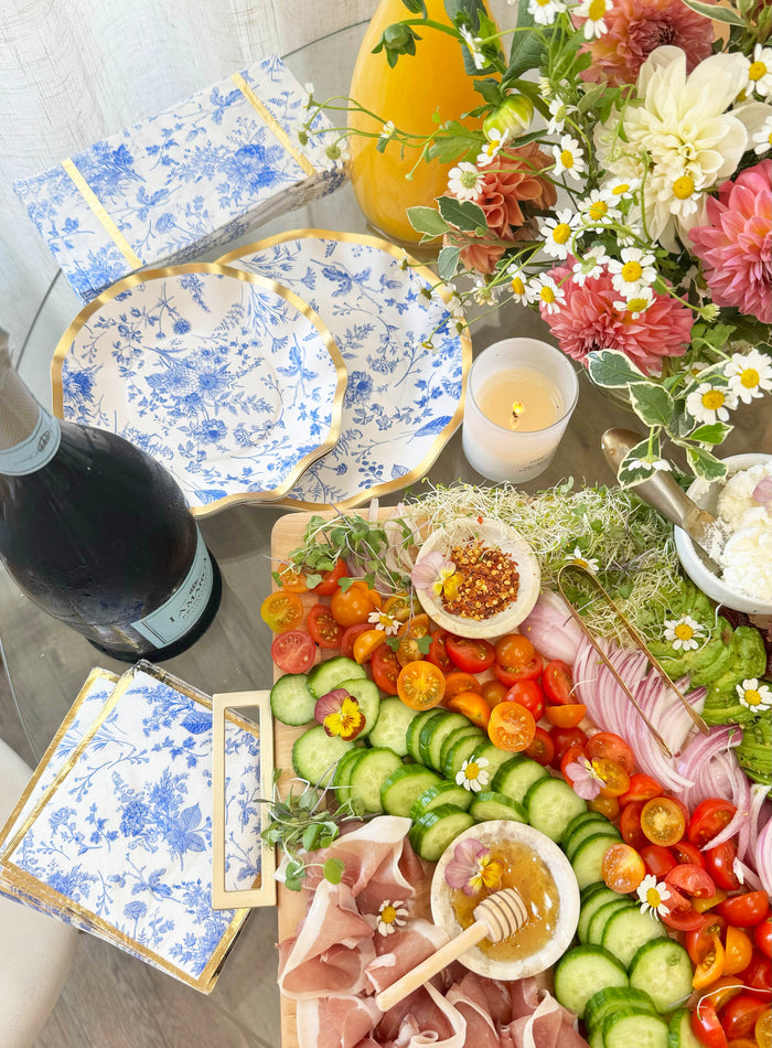 Charcuterie board with prosciutto, cucumber, tomatoes, microgreens, cheese, and honey. Floral napkins, a candle, champagne, flowers, and orange juice are arranged on a glass table alongside Sophistiplate Timeless Dinner Plates (8pk)—perfect for a festive holiday party.