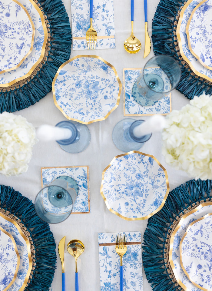 An elegant table setting featuring blue and white floral plates, gold-edged utensils, blue glasses, white flowers, and blue placemats on a crisp white tablecloth is beautifully complemented by Sophistiplate Timeless Guest Towels (20pk), artfully arranged at each place setting and viewed from above.