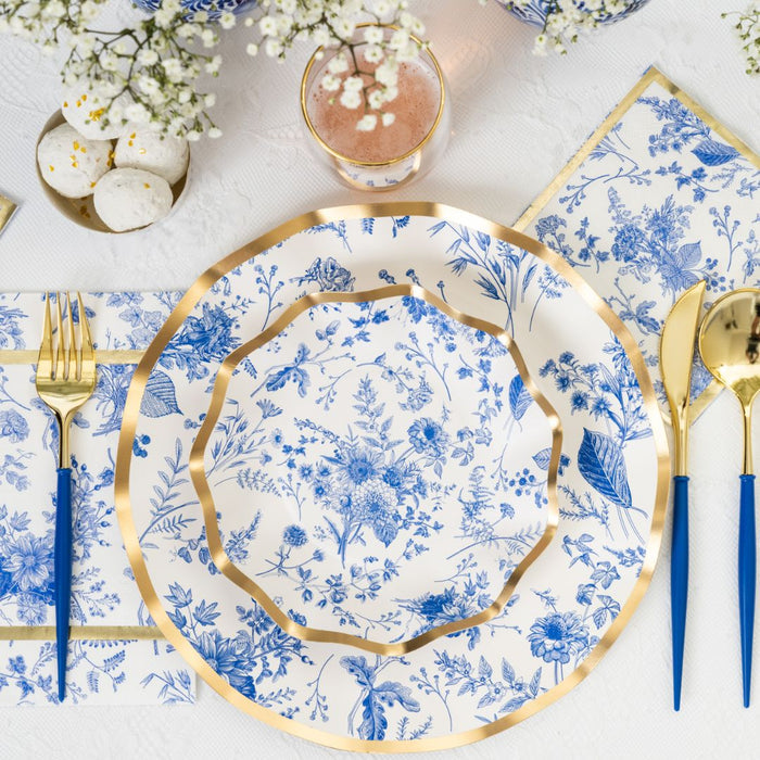 A table setting featuring blue and white floral plates with gold trim, gold and blue cutlery, white macarons, a glass of pink drink, elegant Sophistiplate Timeless Guest Towels, and white flowers on a crisp white tablecloth.