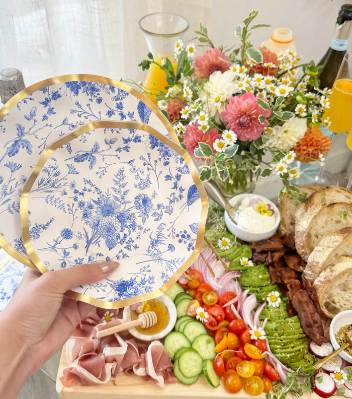 A hand holds two Sophistiplate Timeless Salad Plates from the 8-pack set, each featuring an elegant blue-and-white floral design, above a brunch spread with toast, vegetables, prosciutto, flowers, and drinks on the table.