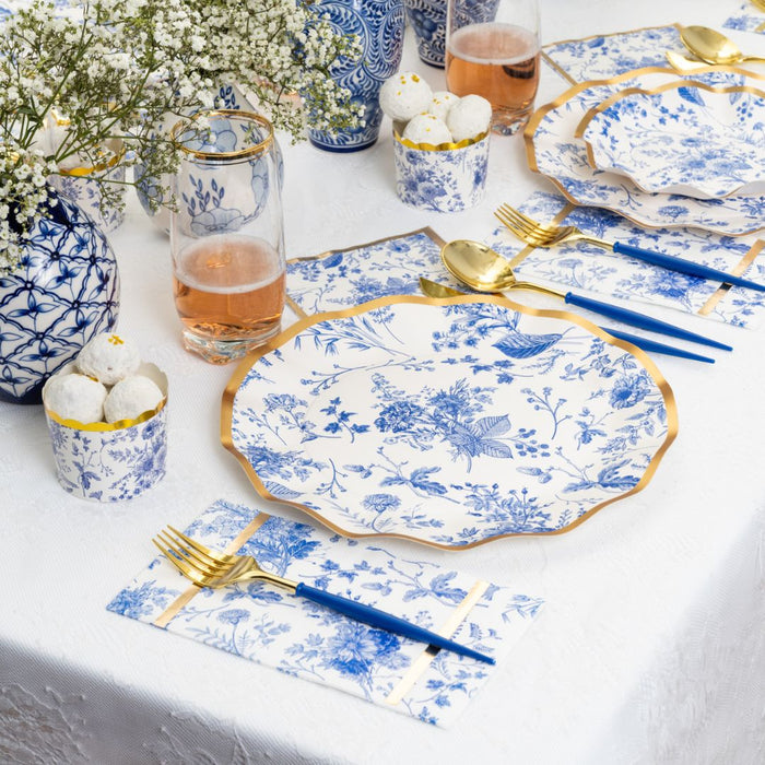 Elevate any sophisticated gathering with the Timeless Salad Plates/8pk by Sophistiplate. The elegant table is set with these blue and white floral gold rim plates, complemented by gold and blue cutlery, glasses of pink drinks, and white flowers in vases atop a crisp white tablecloth. Small bowls filled with delicate white treats complete the look, making these stylish disposable plates an ideal choice for refined events.