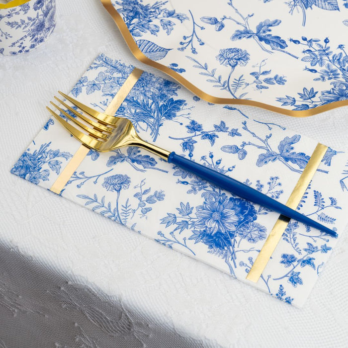 A gold and blue fork rests on Sophistiplate’s Timeless Guest Towels, featuring an elegant blue floral design with gold stripes, next to a blue and white plate with a gold rim, all atop a white lace tablecloth.