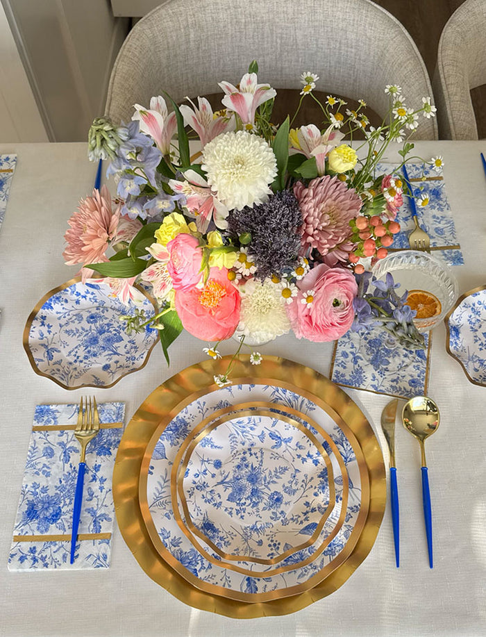 A festive table setting featuring Sophistiplate Timeless Dinner Plates (8pk) with a metallic gold rim and blue floral design, complemented by blue-handled cutlery, coordinated napkins, and a vibrant centerpiece of pink, white, and yellow flowers on a crisp white tablecloth—perfect for creating a memorable holiday party.