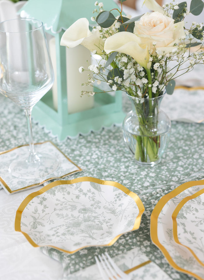 A close-up of an elegant table setting with gold-trimmed floral plates, a wine glass, silverware, matching coasters, and white and cream flowers on a green-patterned runner. Sophistiplate's Timeless Sage Bowls (8pk) add a charming touch alongside the mint lantern.