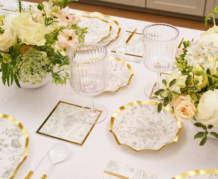 A close-up of an elegant table set with Sophistiplate Timeless Sage Appetizer/Dessert Bowls (8pk), floral-themed plates with gold edges, matching napkins, clear ribbed glasses, white utensils, and floral centerpieces on a white tablecloth.