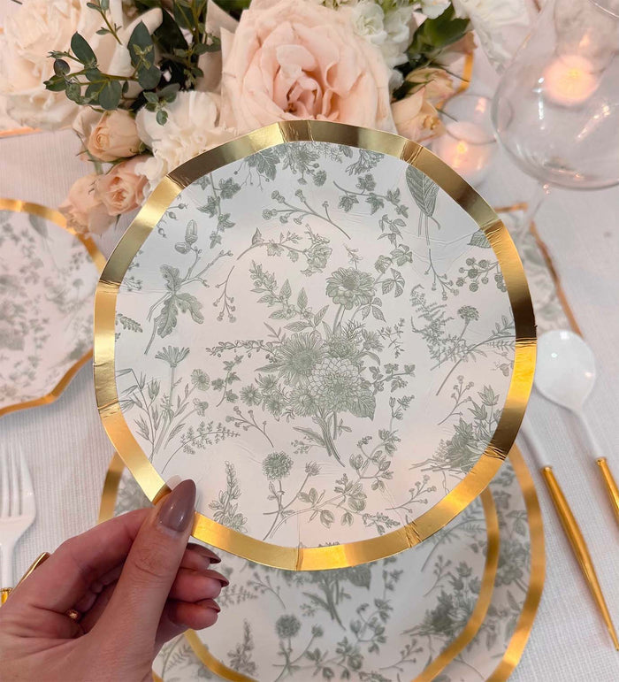 A hand holds a Sophistiplate Timeless Sage Appetizer/Dessert Bowl (8pk) with a gold rim and delicate floral design. In the background are matching scalloped-edge bowls, white utensils, gold straws, candles, and pale flowers on the table.