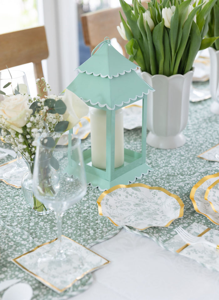 A close-up of a spring table set with a mint green lantern centerpiece, white flowers, wine glasses, gold-trimmed plates, and Sophistiplate Timeless Sage Appetizer/Dessert Bowls/8pk adding charm atop the green floral tablecloth.