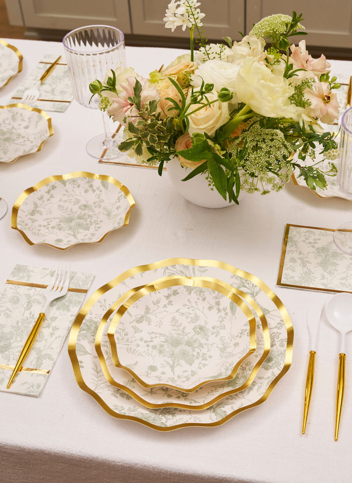 A table set with floral plates edged in gold, matching napkins, clear cups, and gold cutlery is complemented by Sophistiplate Timeless Sage Appetizer/Dessert Bowls (8pk) and a white vase with white and pale pink flowers in the center.